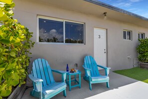 A building with a door marked with the number 3, two blue Adirondack chairs, a small table with a bottle and fruit, and a window with blinds.