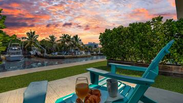 A table with a glass of beverage, a mug, and a plate with food, overlooking a pool with boats and palm trees under a sunset sky.