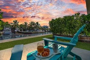Une table avec un verre contenant une boisson, une tasse et une assiette remplie de nourriture, donnant sur une piscine où flottent des bateaux, avec des palmiers se découpant sur un ciel au coucher du soleil.