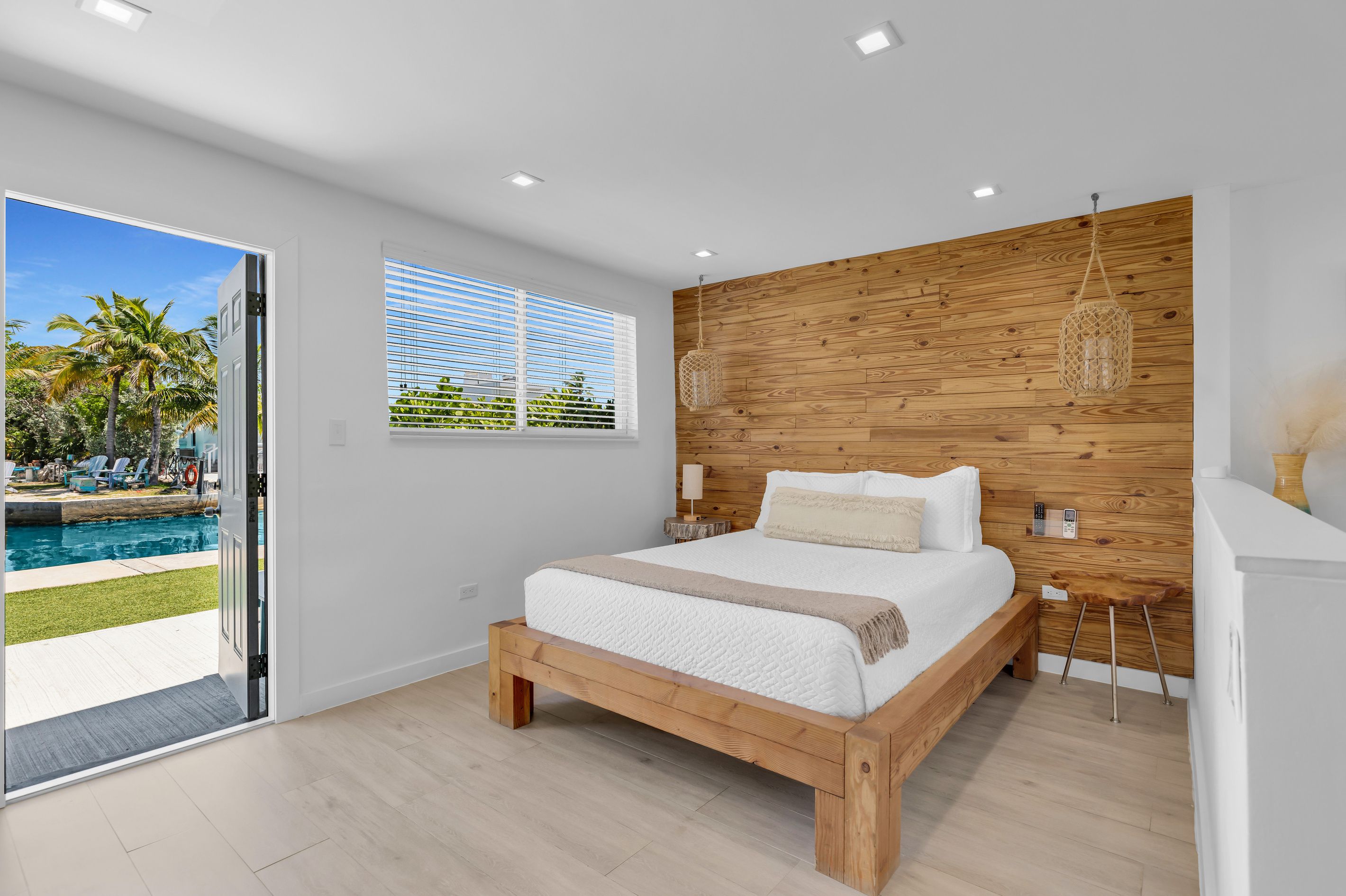Ein Schlafzimmer mit einem Holz-Kopfteil, einem großen Bett, einem Nachttisch mit Lampe und Blick auf einen Pool im Freien.