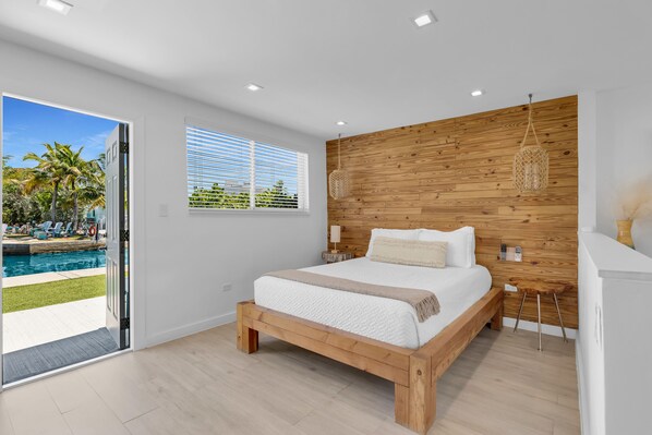 A bedroom with a wooden headboard, a large bed, a bedside table with a lamp, and a view of a swimming pool outside.