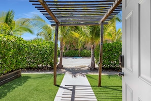 A covered outdoor area with a pathway leading to a pool, surrounded by lush greenery and palm trees.