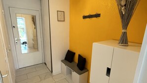 A hallway with a white door, a bench with black cushions, a wall-mounted guitar, and a vase with dried plants.