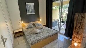 A bedroom with a large bed, two bedside tables, a nightstand, a wall-mounted light, and a view of the outdoors through a sliding glass door.