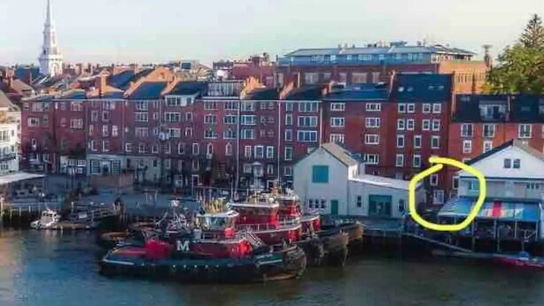 Stadsgezicht aan het water met rode bakstenen gebouwen, een steiger met boten, en een opvallende toren op de achtergrond.