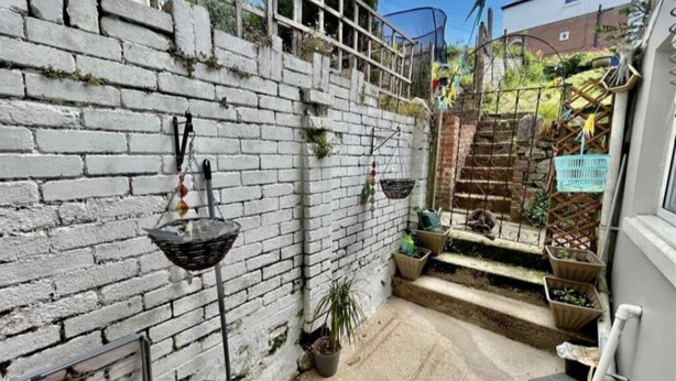 Un espacio exterior reducido con un muro de ladrillo blanco, una planta pequeña en maceta y una jardinera colgante de metal.