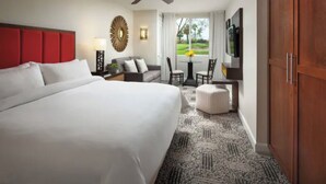 Quarto de hotel com uma cama grande, cabeceira vermelha, sofá, pequena mesa com cadeiras e televisão na parede.