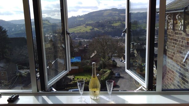 Une bouteille de champagne et deux verres sont posés sur un rebord de fenêtre, d’où l’on aperçoit une ville et des collines.