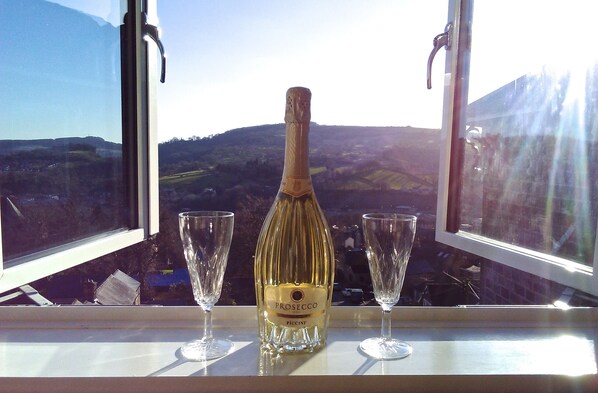 Una botella de Prosecco y dos copas sobre una mesa junto a una ventana con vistas al paisaje.