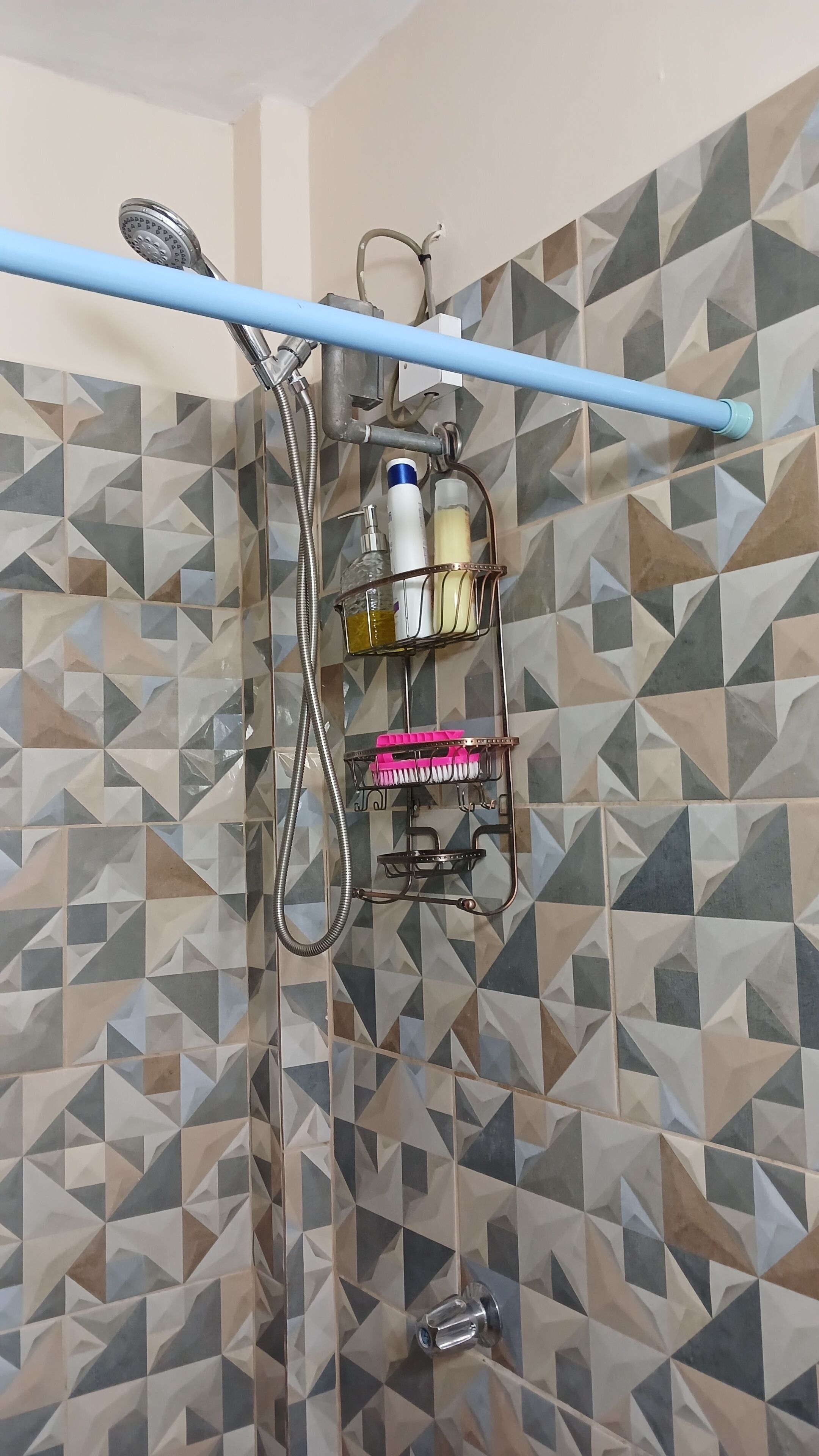 Zona de ducha con un diseño de azulejos geométrico, un cabezal de ducha manual, una manguera azul y un estante metálico con artículos de aseo.