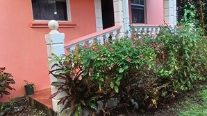 A house with a pink facade, white porch, and a garden with flowering plants.