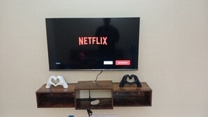A television displaying the Netflix logo, with a wooden shelf below featuring two heart-shaped objects and a small rock.