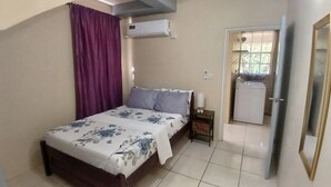 A bedroom with a bed, a nightstand, a window with curtains, an air conditioning unit, and a door leading to a laundry area.
