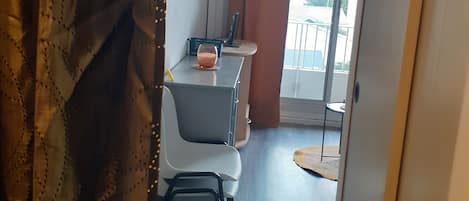A room with a desk, chair, and a small table with a glass of drink. There is a window with curtains and a view of the outside.