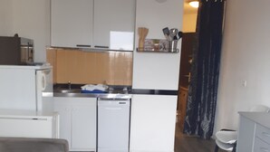 A compact kitchen with white cabinets, a built-in oven, and a microwave. There's a small round table with a stool in the living area, and a grey sofa. - Pleasant T1 hyper center Valras plage (Valras-Plage)