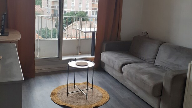 A living room with a grey sofa, a small round table, and a view of buildings through a window with brown curtains. - Pleasant T1 hyper center Valras plage (Valras-Plage)