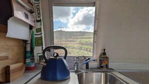 Een keuken met een blauwe waterkoker op een mat, een roestvrijstalen spoelbak en een raam met uitzicht op een heuvel.