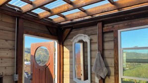 Een houten badkamer met een houten deur, een ronde spiegel, een witte wastafel en een raam met uitzicht op de natuur.