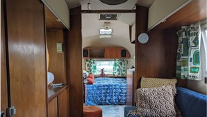 Een compact interieur van een camper, met een kitchenette, een bed met een blauw beddegoed met patroon en een blauwe bank met geknoopte kussens.