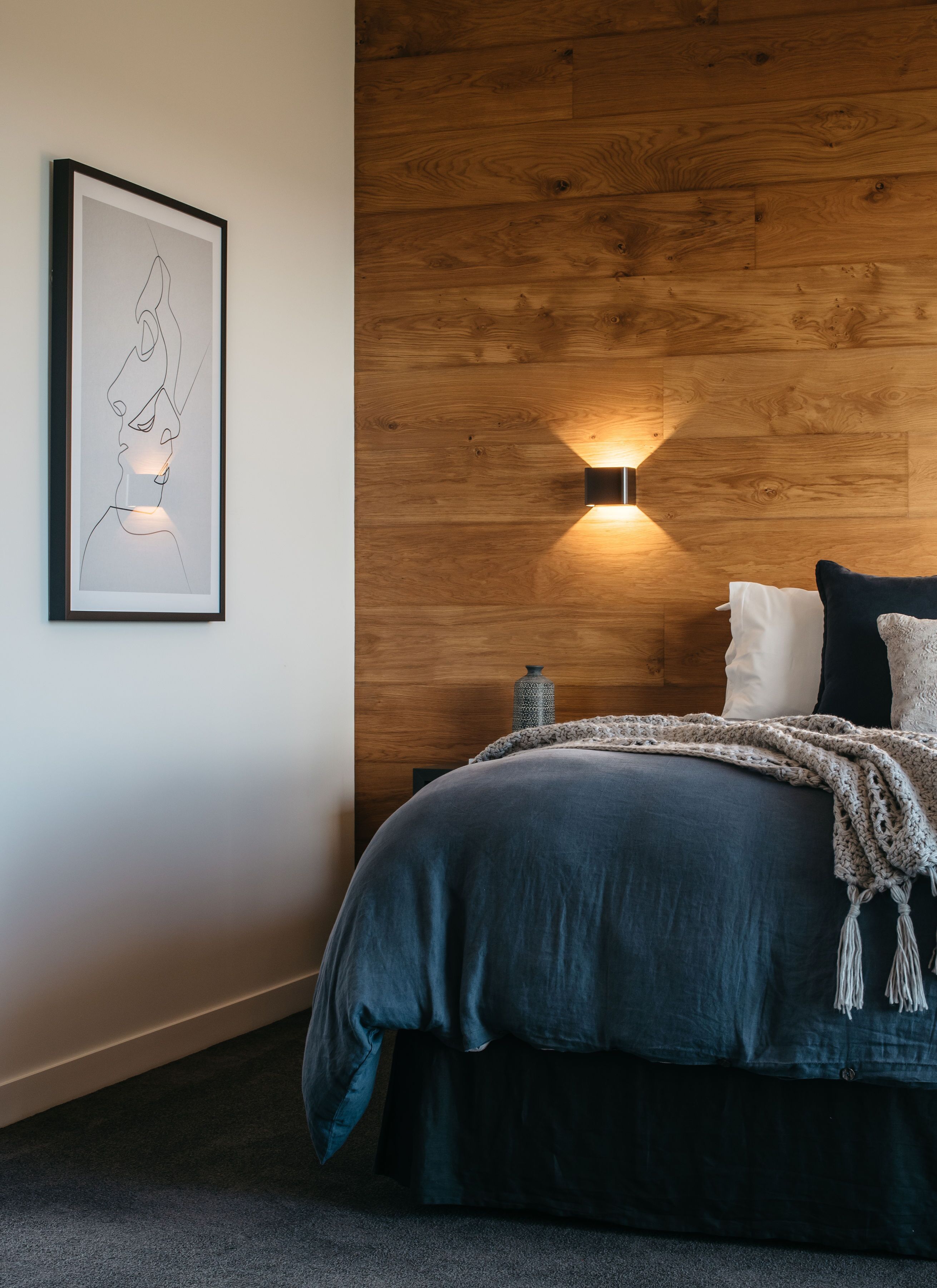 A bedroom with a wooden headboard, a framed artwork, a bedside lamp, and a bed with a dark blue bedspread and pillows.