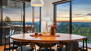 A modern dining area with a round table, black chairs, and a view of a sunset over a forested landscape through large windows. - The Ridge at Maleny (Booroobin)
