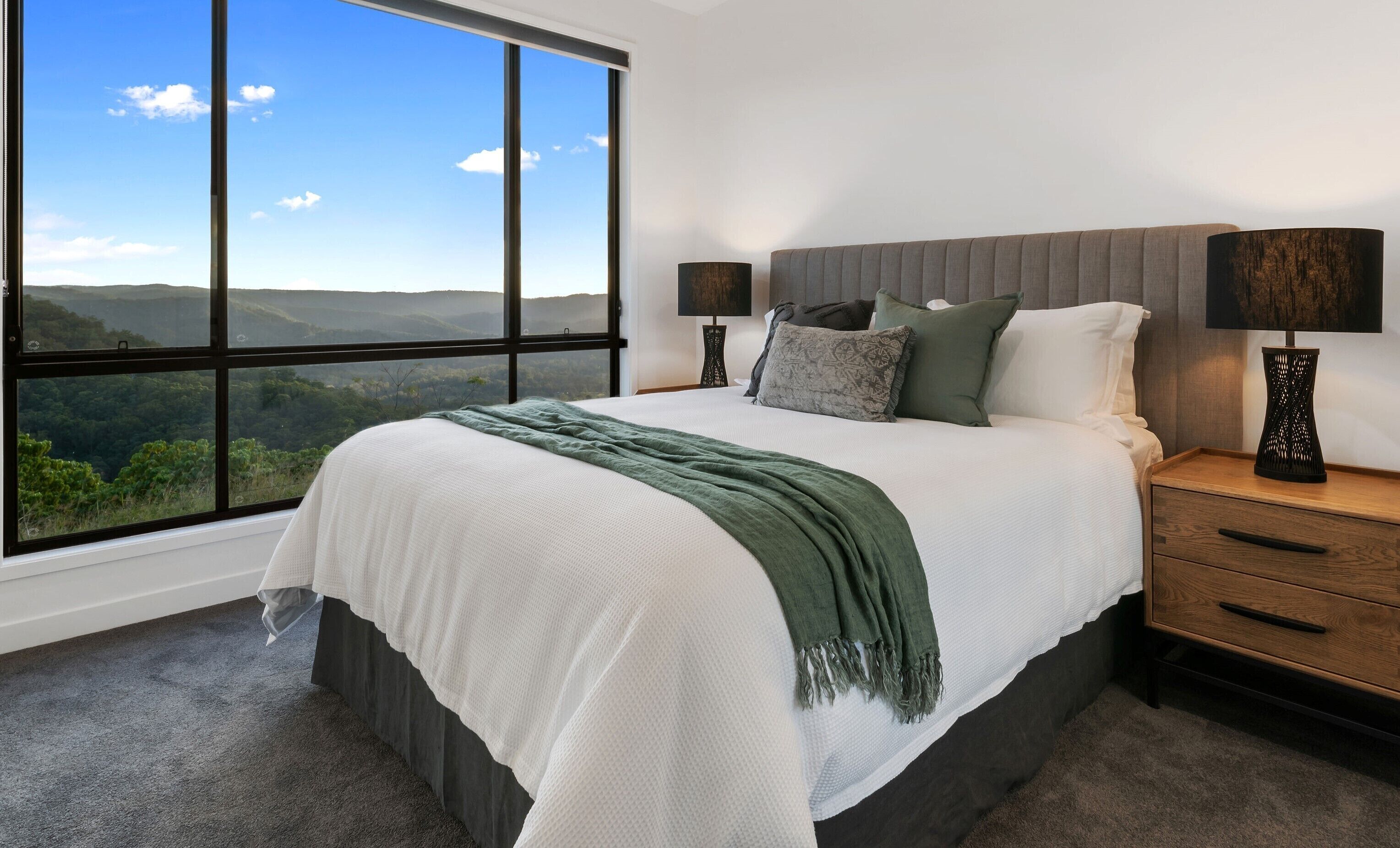 A bedroom with a large window, a bed with a green throw, two bedside lamps, and a nightstand with a drawer.