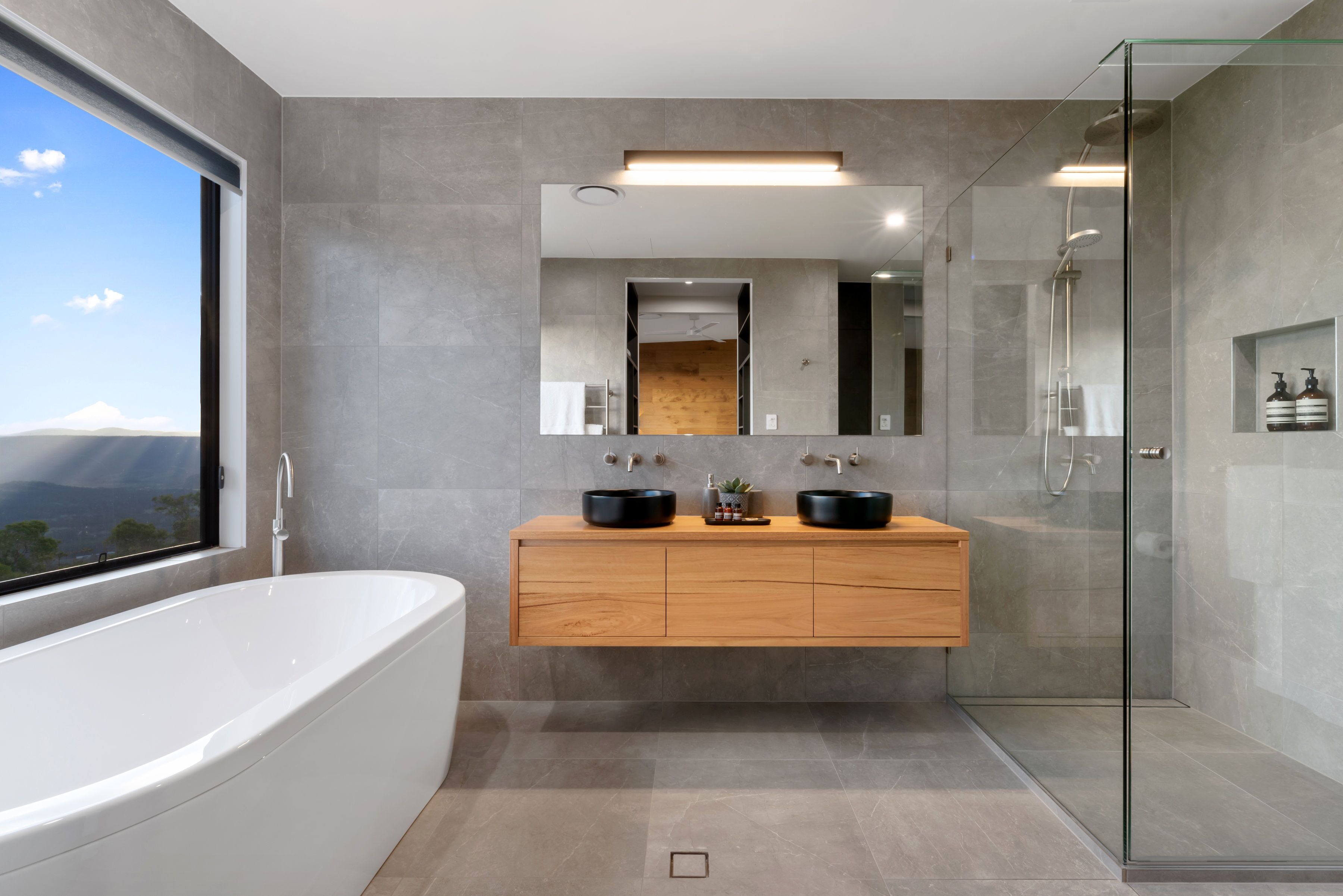 A modern bathroom with a freestanding bathtub, a wooden vanity with two black sinks, a glass shower enclosure, and a large mirror above the vanity.