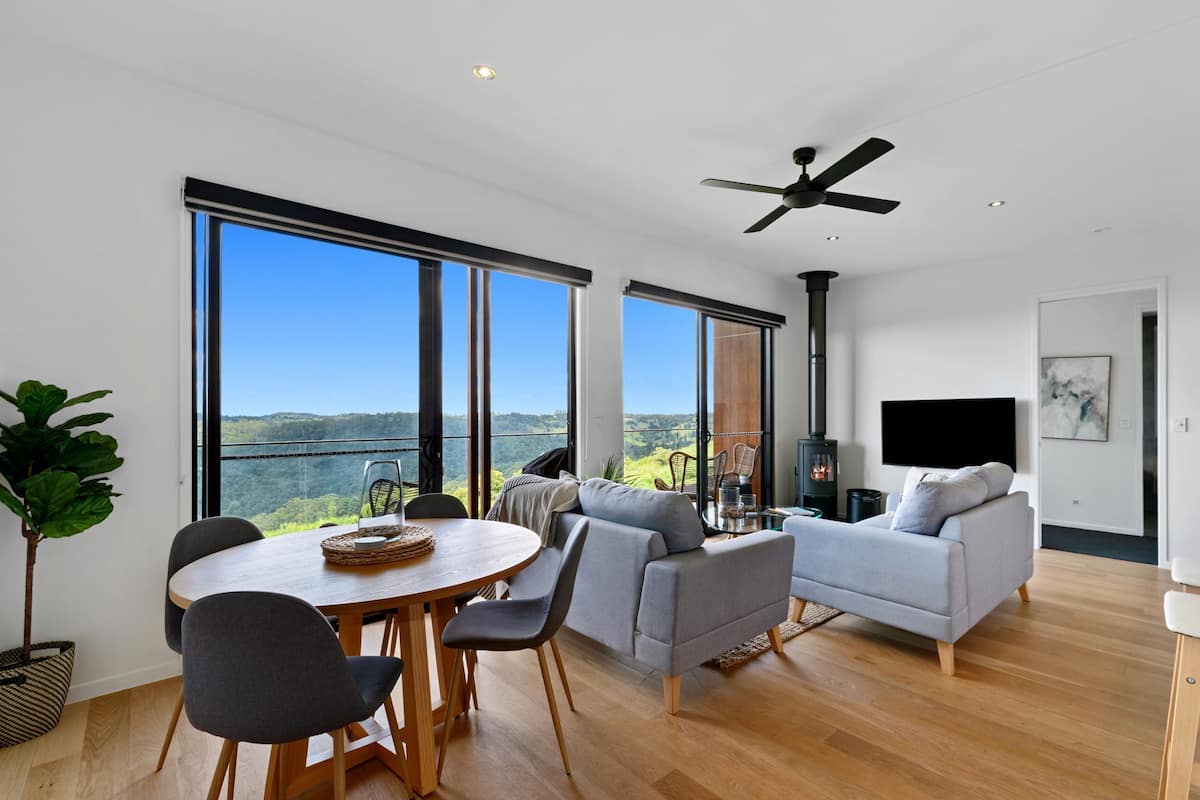 A spacious living area with a large window, a ceiling fan, a fireplace, and a flat-screen TV.