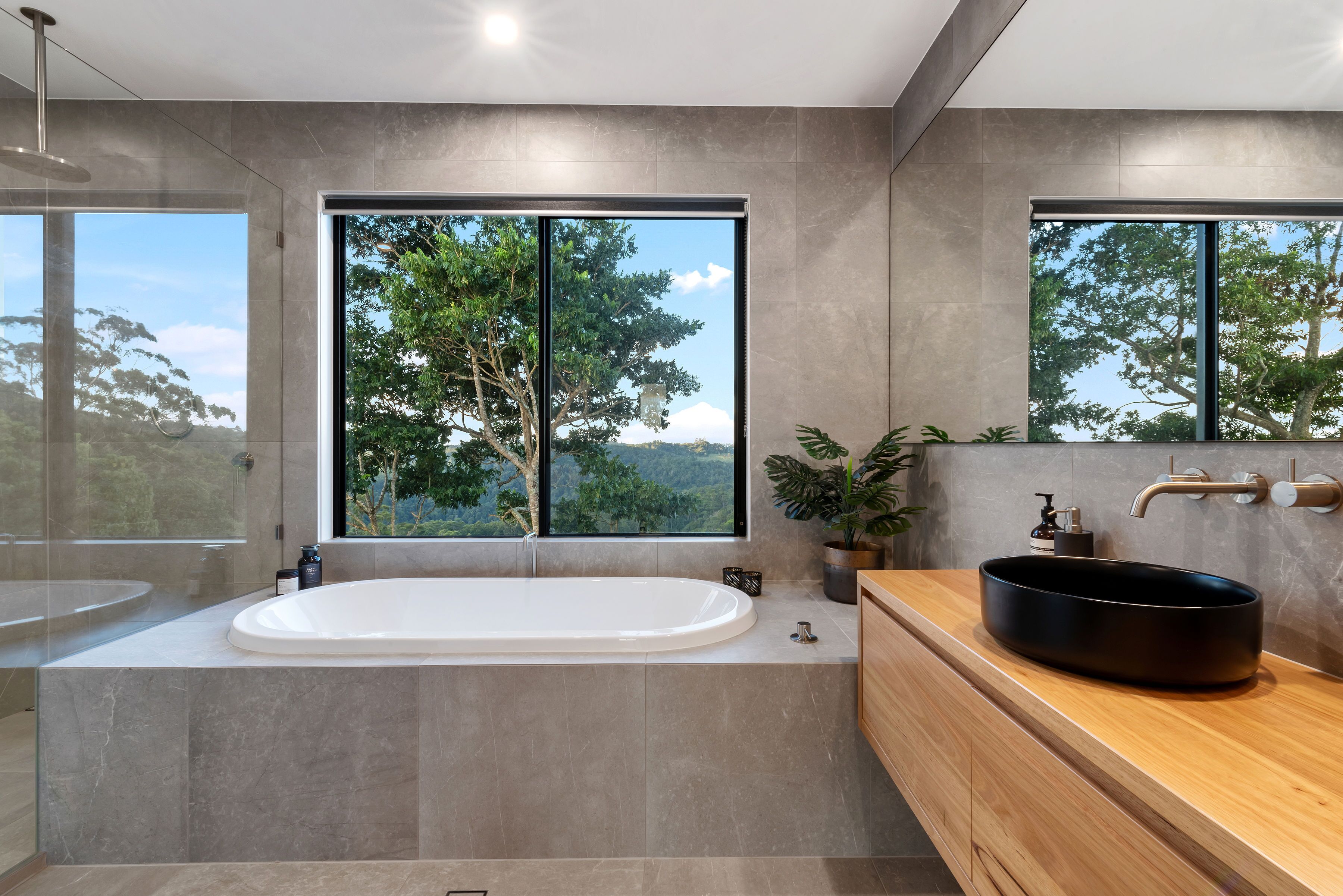 A modern bathroom with a freestanding bathtub, a wooden vanity, and a large window offering a view of trees and hills.
