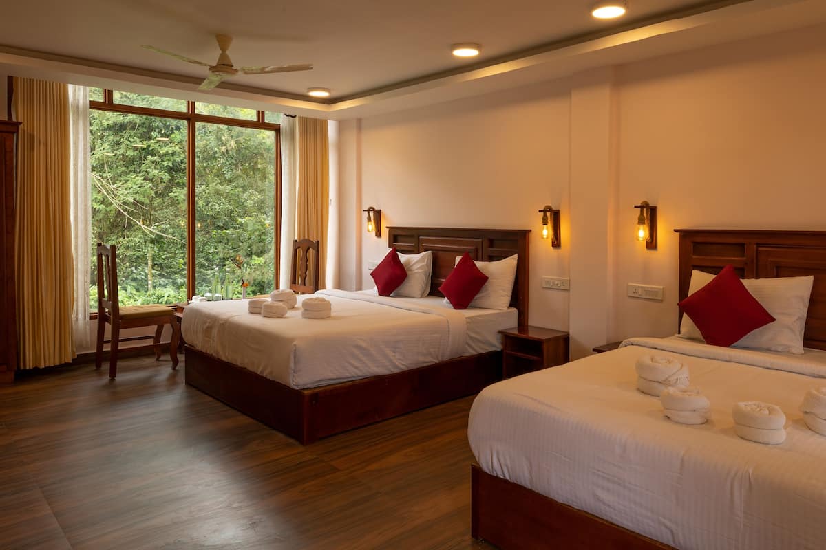 A hotel room with two beds, wooden flooring, a large window with a view of greenery, and a ceiling fan.
