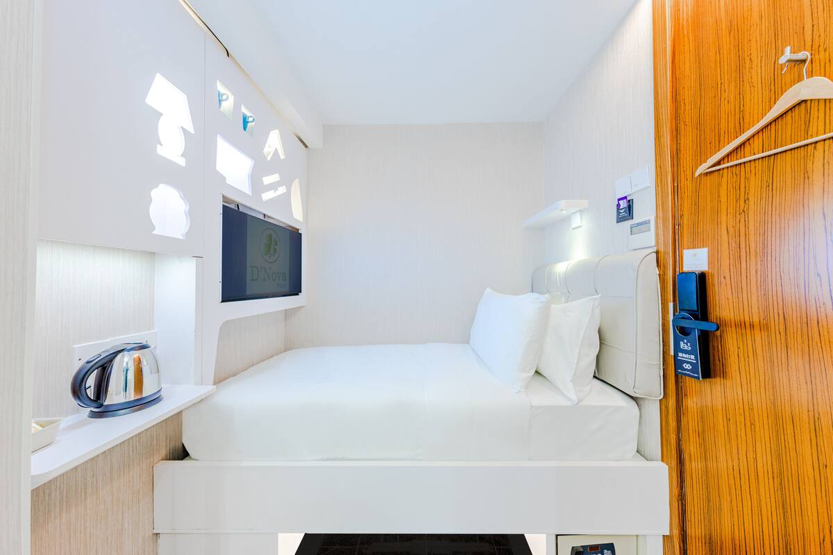 A compact hotel room with a bed, a bedside table with a kettle, a wall-mounted TV, and a wooden door.