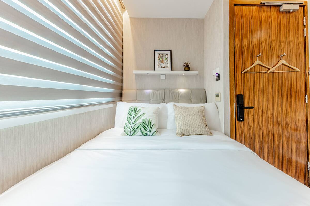 A bedroom with a bed, a wooden door, a shelf with a picture and a plant, and a window with blinds.