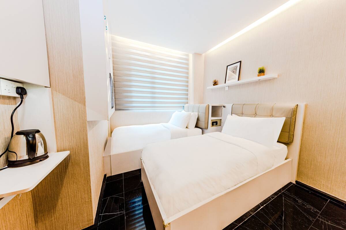 A modern hotel room with a large bed, a smaller sofa, a kettle on a shelf, and a window with blinds.