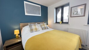 A neatly made bed with a yellow blanket, a wooden headboard, and two plaid pillows. There is a bedside lamp, a window with blinds, and a radiator below. - 6 Cross Court, Stafford (Stafford)