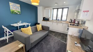 A compact living space with a grey sofa, a small dining table, and a kitchenette. - 6 Cross Court, Stafford (Stafford)