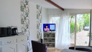 A room with a wooden floor, white furniture, a television, and a view of greenery outside. - Meublé de tourisme classé 3* 35 m2 + terrasse. 2 pers, countryside VEXIN FRANçAIS (Guernes)