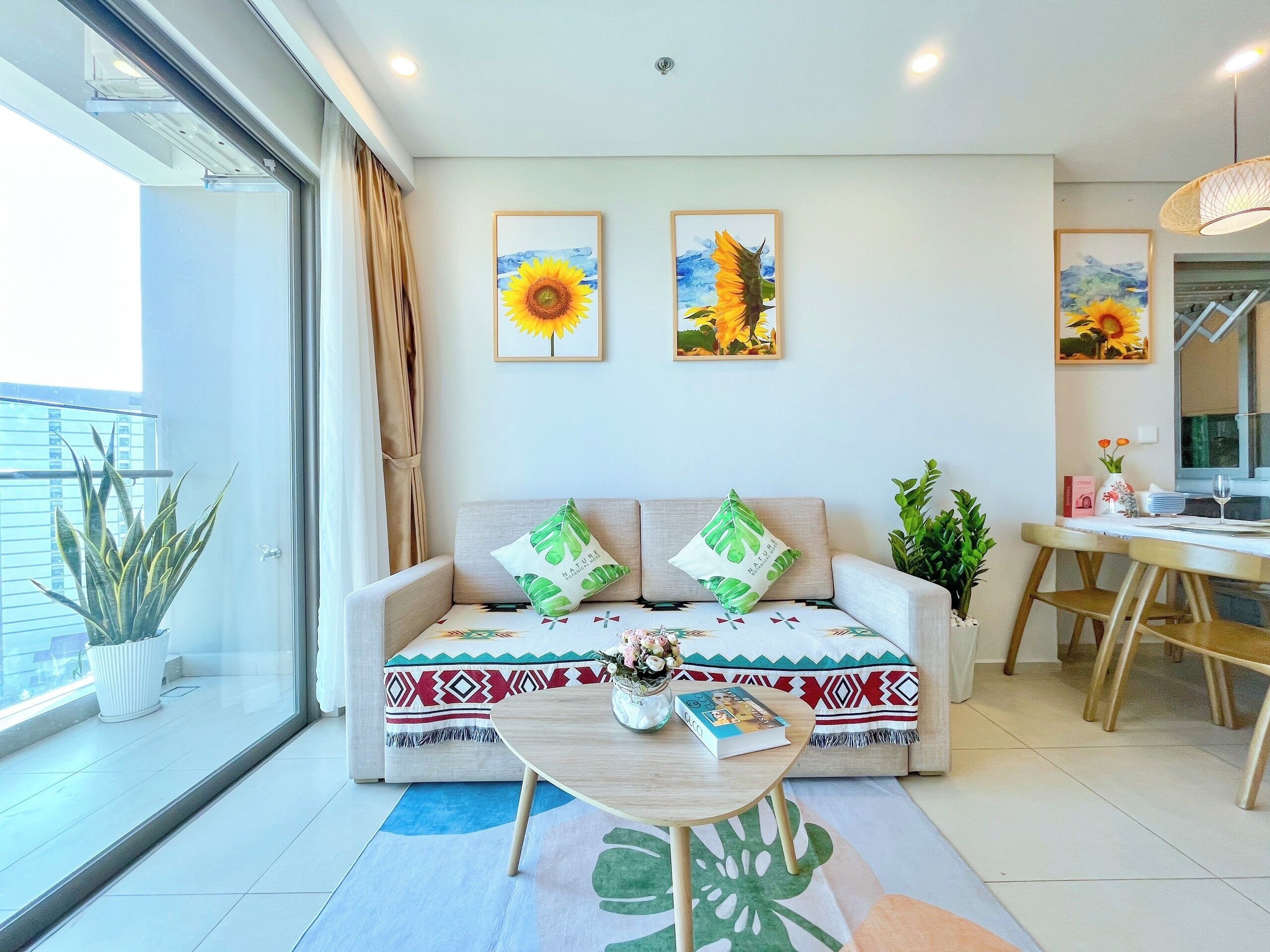 Ein modernes Wohnzimmer mit einer Couch, einem Couchtisch und einem Essbereich. An der Wand hängen zwei gerahmte Kunstwerke, eine Topfpflanze und es gibt einen Blick auf den Balkon.