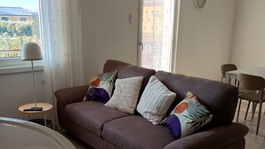 A living room with a brown sofa, a small round table, and a large window with curtains. - Casa Viola: Your Relaxing Retreat by the Lake (Pacengo)
