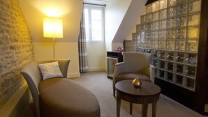 A cozy room with a sofa, armchair, and a small table. There is a lamp, a window with curtains, and a decorative glass wall. - Hotel The Originals de la Paix Beaune (Beaune)