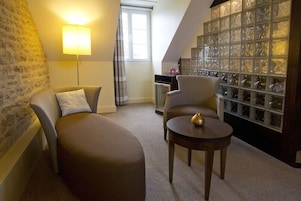 Una habitación acogedora con sofá, sillón y una pequeña mesa. Hay una lámpara, un ventanal con cortinas y una pared decorativa de vidrio.