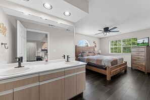 A modern bedroom with a large bed, a vanity with dual sinks, a ceiling fan, and a view of greenery through the window.