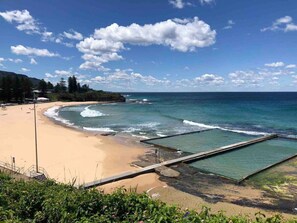 A beach with clear skies, calm sea, and a rocky outcrop with a viewing platform.