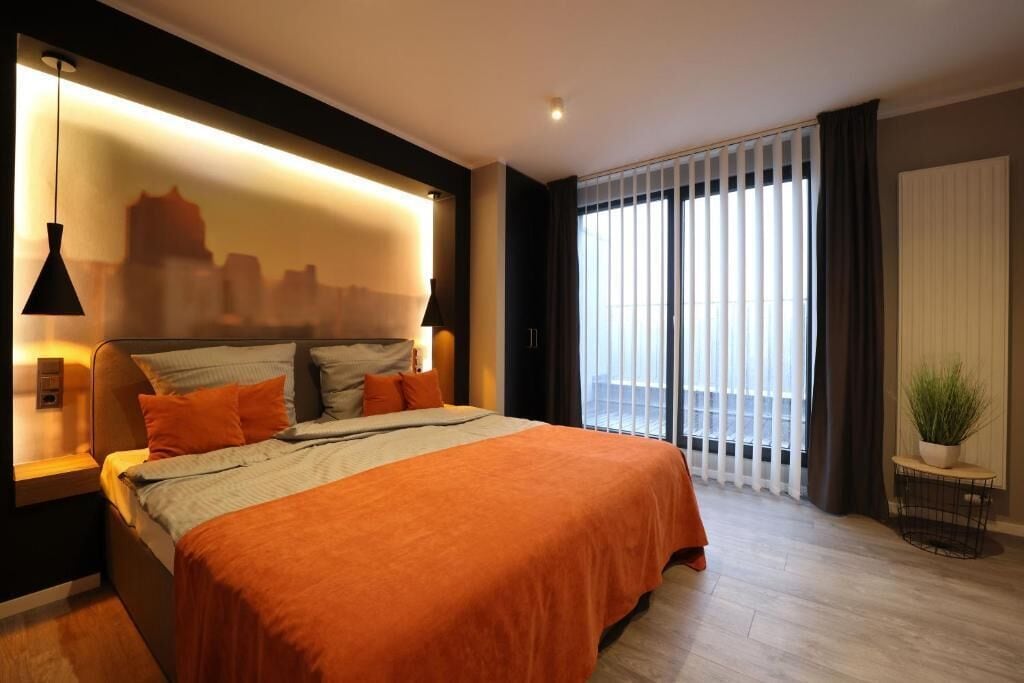 A modern bedroom with a large bed, a cityscape mural on the wall, a window with blinds, and a small plant on a side table.