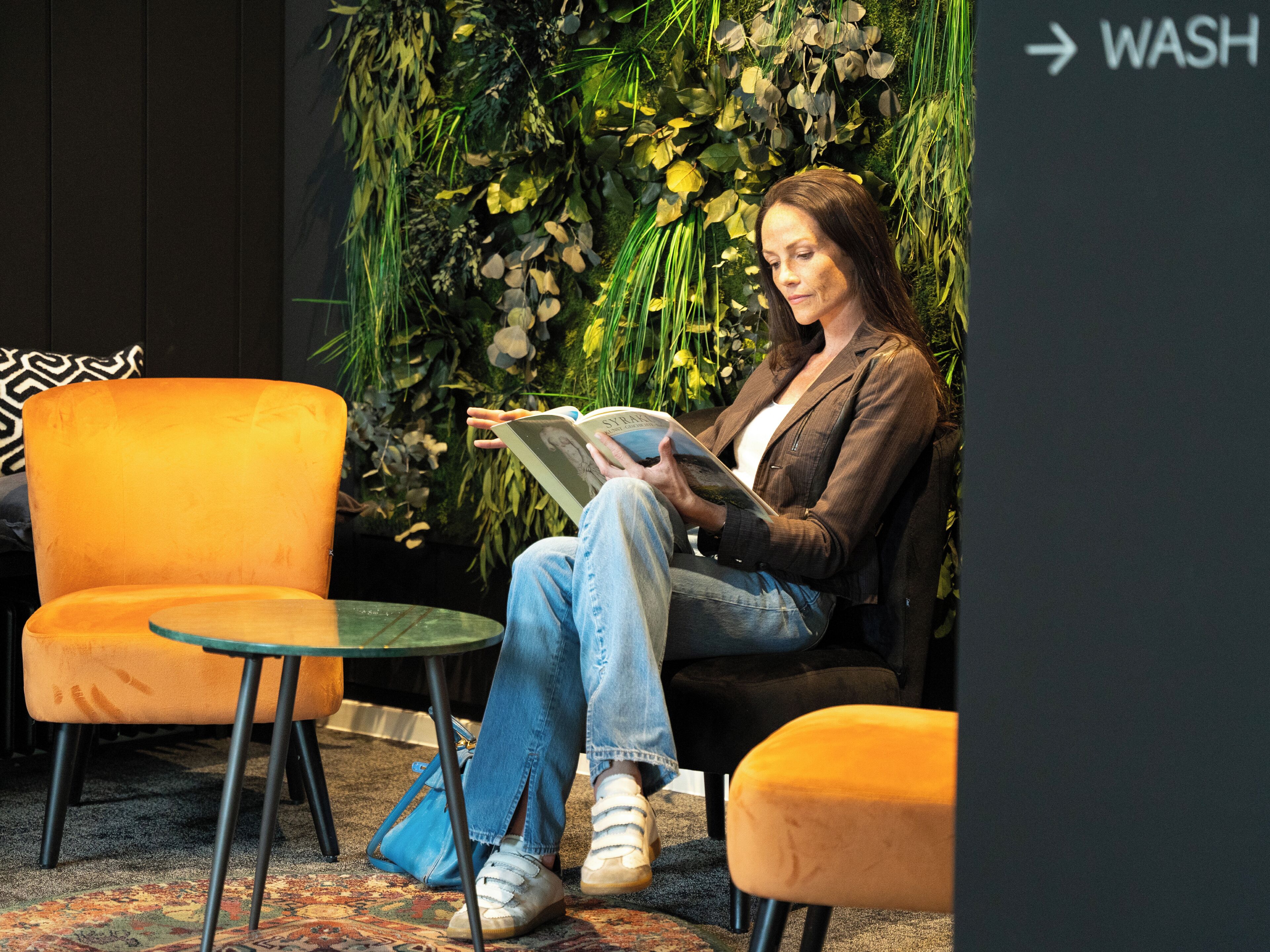 Una persona sentada en una silla naranja leyendo un libro en una habitación con una pared cubierta de hojas verdes y un letrero negro que dice 'WASH' y una flecha.