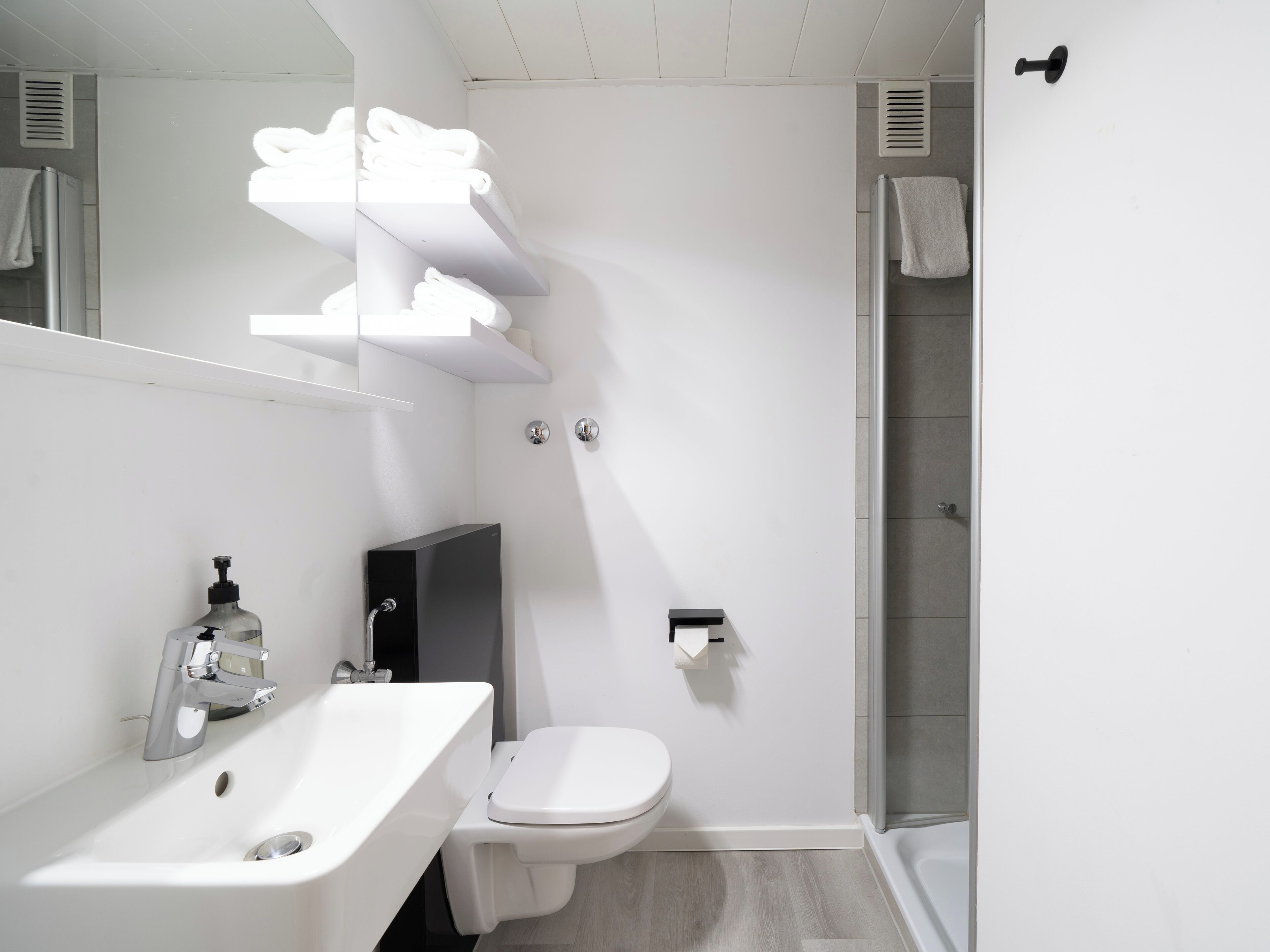Un baño moderno con lavamanos, inodoro y ducha blancos. Hay repisas sobre el lavamanos con toallas.