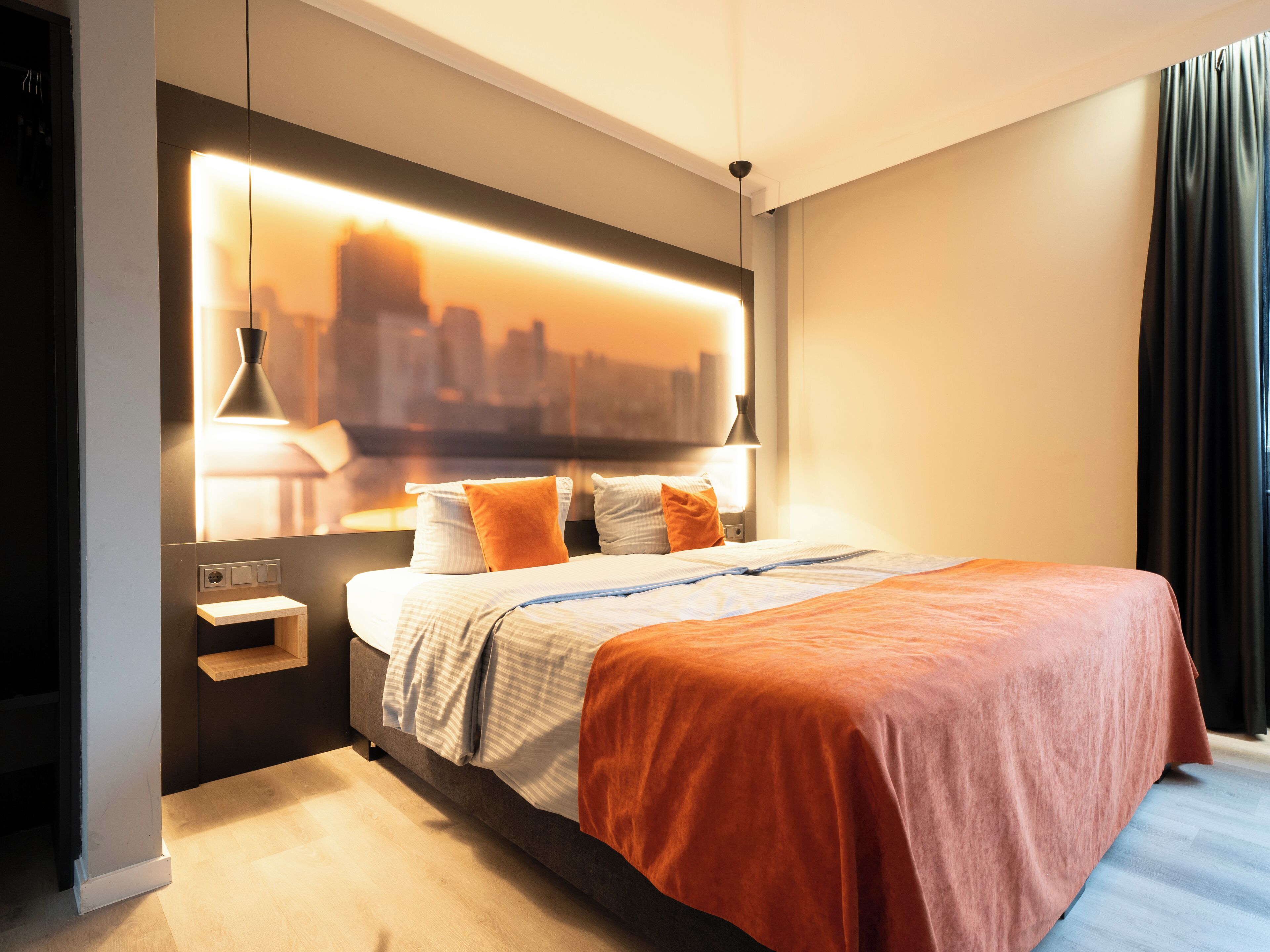 A modern hotel room with a large bed, city skyline view, and a bedside table with lamps.