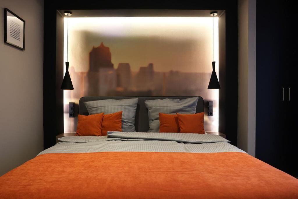 Une chambre d’hôtel moderne avec vue sur la ville, un lit bien fait avec une literie orange et deux lampes suspendues noires.