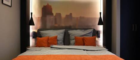 Une chambre d’hôtel moderne avec vue sur la ville, un lit bien fait avec une literie orange et deux lampes suspendues noires.