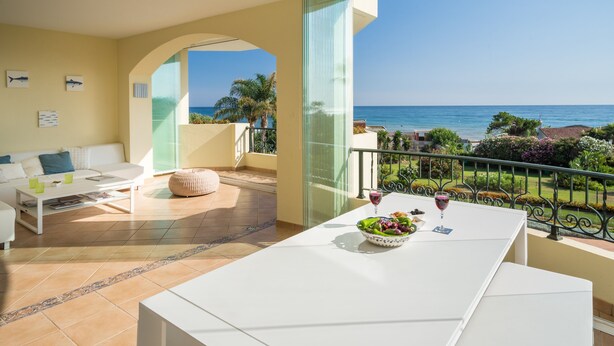 A modern living room with a balcony overlooking the sea, a dining table with a bowl of fruit and wine glasses, and a white sofa. - Wow☀️☀️☀️☀️☀️ Beachfront apartment, panoramic views, amazing sunsets, XL pool! (Marbella)