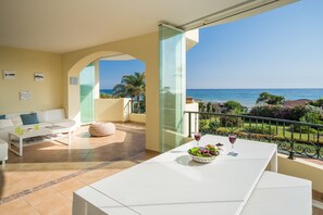 Uma sala de estar moderna com varanda e vista para o mar, uma mesa de jantar com uma tigela de frutas e copos de vinho, e um sofá branco.
